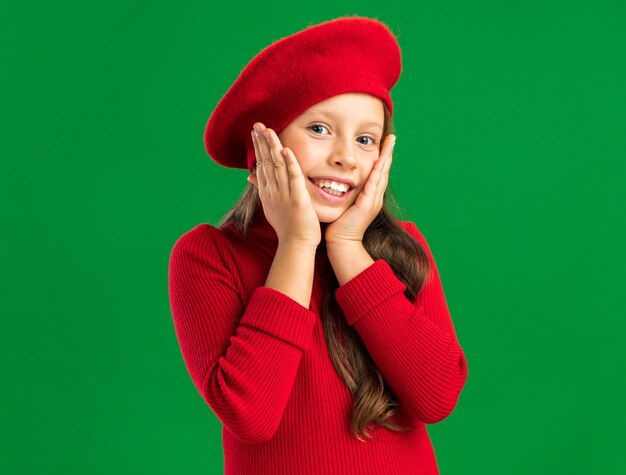 Radosna mała blondynka w czerwonym berecie trzymająca ręce na twarzy na zielonej ścianie z kopią miejsca