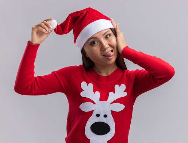 Radosna młoda azjatycka dziewczyna ubrana w świąteczny kapelusz ze swetrem pokazując język na białym tle