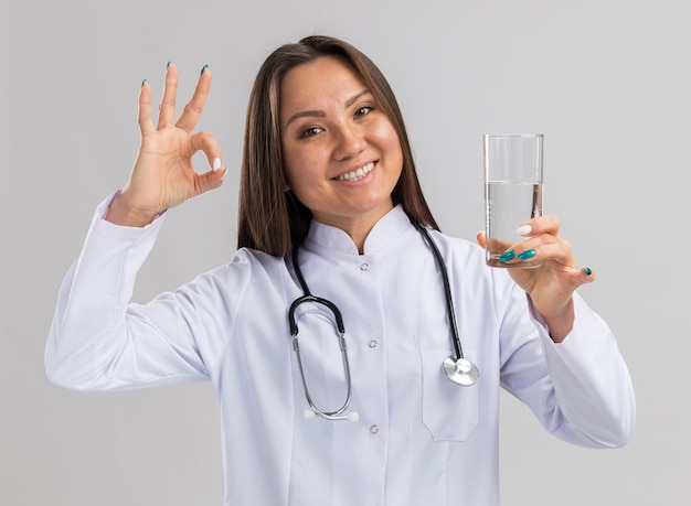Radosna młoda azjatycka lekarka nosi szatę medyczną i stetoskop trzymając szklankę wody patrząc na przód robi ok znak na białym tle na białej ścianie