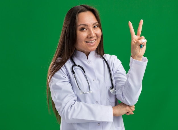 Radosna młoda azjatycka lekarka nosząca medyczną szatę i stetoskop, patrząc na przód robi znak pokoju na zielonej ścianie z kopią przestrzeni