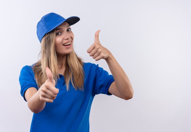 Radosna młoda dziewczyna dostawy ubrana w niebieski mundur i czapkę kciuki do góry na białym tle na białej ścianie z miejsca na kopię