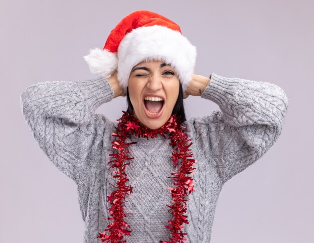 Radosna młoda kaukaska dziewczyna ubrana w świąteczny kapelusz i świecącą girlandę na szyi, trzymając ręce na głowie, krzycząc i mrugając na białym tle na białej ścianie