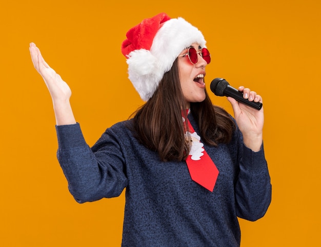 Bezpłatne zdjęcie radosna młoda kaukaska dziewczyna w okularach przeciwsłonecznych z czapką świętego mikołaja i krawatem świętego mikołaja trzyma mikrofon, udając, że śpiewa, patrząc na stronę odizolowaną na pomarańczowej ścianie z kopią przestrzeni