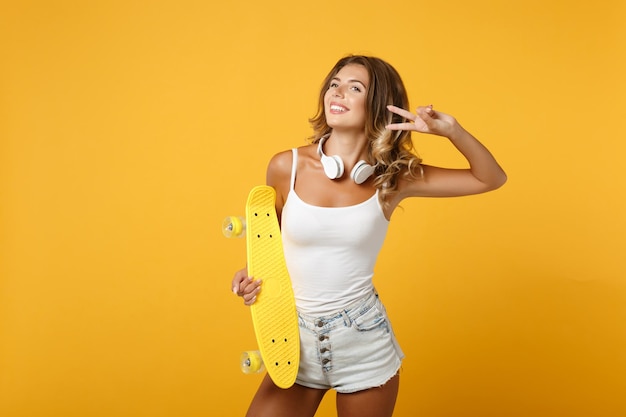 Radosna młoda kobieta dziewczyna w lekkim ubraniu ze słuchawkami pozowanie na białym tle na żółtym pomarańczowym tle. koncepcja życia ludzi. makieta miejsca na kopię. trzymając deskorolkę, pokazując znak zwycięstwa.