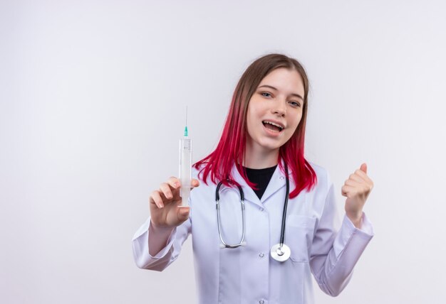 Radosna młoda kobieta lekarz ubrana w stetoskop medyczny szlafrok trzymając strzykawkę podnosząc pięść na odosobnionej białej ścianie
