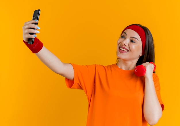 Radosna młoda kobieta sportowy noszenia opaski i nadgarstków, trzymając hantle i biorąc selfie