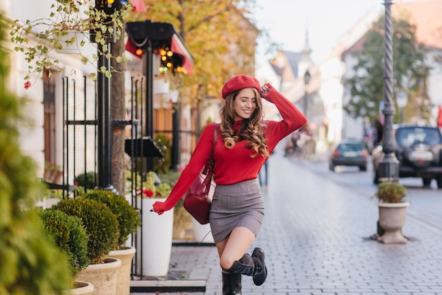 Radosna młoda kobieta w czerwonym berecie tańczy na chodniku z czarującym uśmiechem