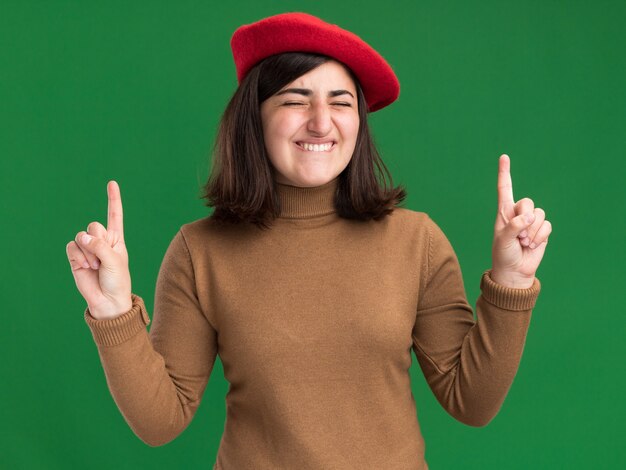 Radosna młoda ładna kaukaska dziewczyna z beretowym kapeluszem stoi z zamkniętymi oczami skierowanymi w górę na zielonej ścianie z kopią przestrzeni