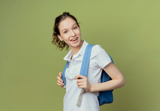 Radosna młoda ładna studentka noszenie plecaka stojącego w widoku profilu trzymając paski torby na białym tle na oliwkowym tle z miejsca kopiowania