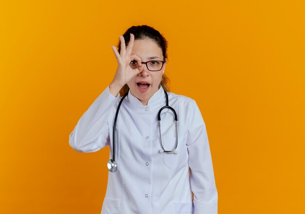 Radosna młoda lekarka na sobie szlafrok medyczny i stetoskop w okularach pokazujących gest wygląd na białym tle