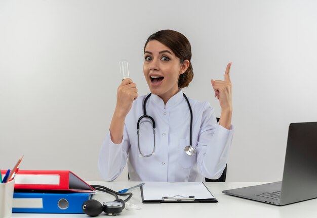 Radosna młoda lekarka ubrana w szlafrok medyczny ze stetoskopem siedząca przy biurku pracuje na komputerze z narzędziami medycznymi trzymając żarówkę kciuk w górę na odizolowanej białej ścianie z miejscem na kopię