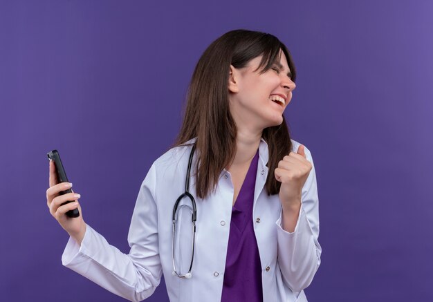 Radosna młoda lekarka w szlafroku medycznym ze stetoskopem trzyma telefon i patrzy w bok na odosobnionym fioletowym tle z miejscem na kopię