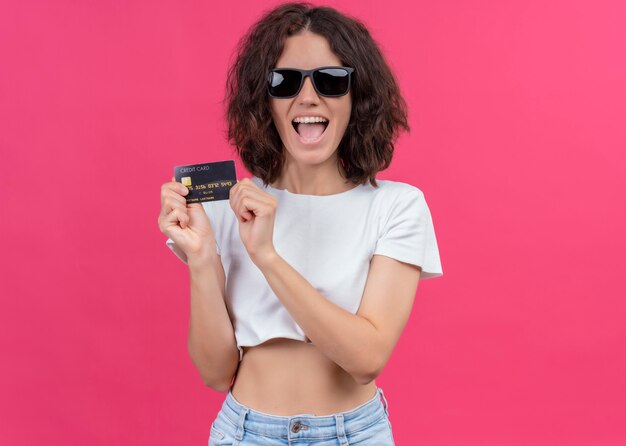 Radosna młoda piękna kobieta nosi okulary przeciwsłoneczne i trzyma kartę na odosobnionej różowej ścianie z miejsca na kopię