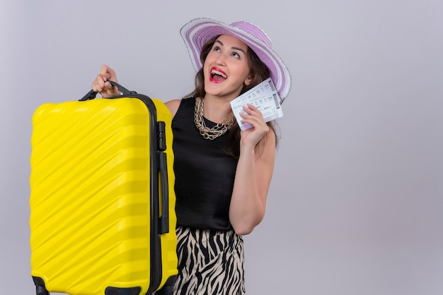 Radosna młoda podróżniczka na sobie czarny podkoszulek w kapeluszu, trzymając walizkę i bilety na białej ścianie