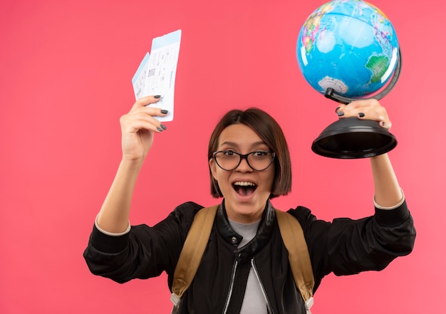 Bezpłatne zdjęcie radosna młoda studentka w okularach i plecak podnosząca bilety lotnicze i kula ziemska na różowym tle