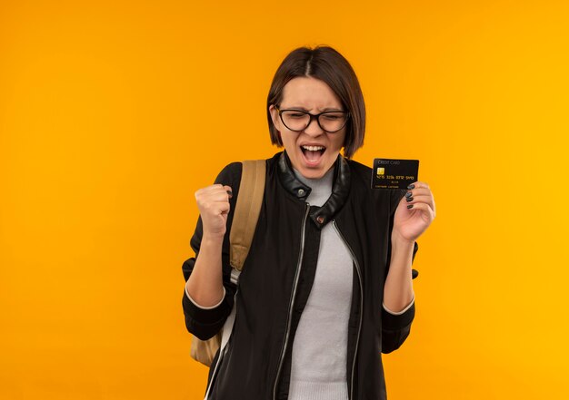 Radosna młoda studentka w okularach iz powrotem worek trzyma kartę kredytową zaciskając pięść z zamkniętymi oczami odizolowane na pomarańczowo