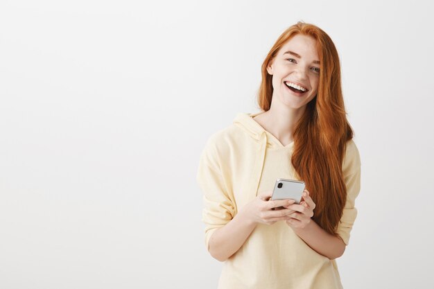 Radosna ruda dziewczyna uśmiechnięta i śmiejąca się, za pomocą smartfona