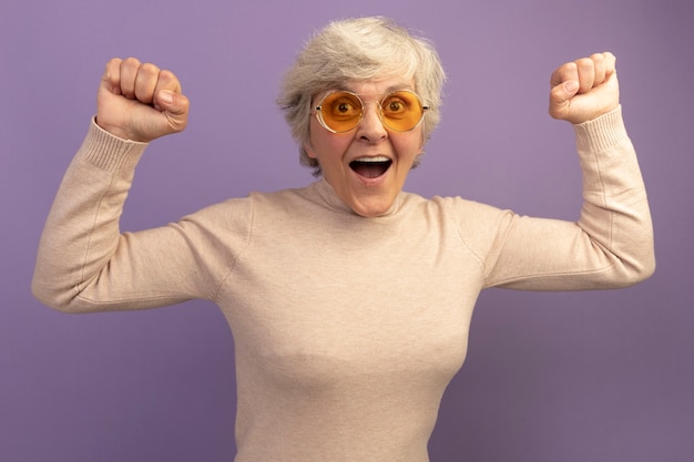 Radosna stara kobieta w kremowym swetrze z golfem i okularach przeciwsłonecznych, patrząca z przodu, wykonująca gest „tak” na fioletowej ścianie