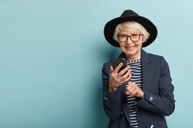 Radosna starsza kobieta ma okulary optyczne, robi bankowość online na telefonie komórkowym, wykorzystuje nowoczesne technologie do wyszukiwania informacji w Internecie, ubrana w modne ciuchy, uśmiechnięta, odizolowana