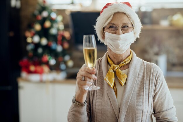 Radosna starsza kobieta z maską na twarz unosząca szklankę podczas świętowania Nowego Roku w domu