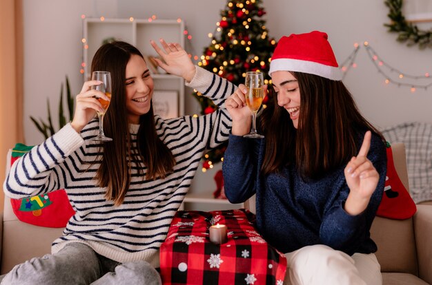 Radosne ładne młode dziewczyny trzymają kieliszki szampana i tańczą, siedząc na fotelach i ciesząc się świętami Bożego Narodzenia w domu