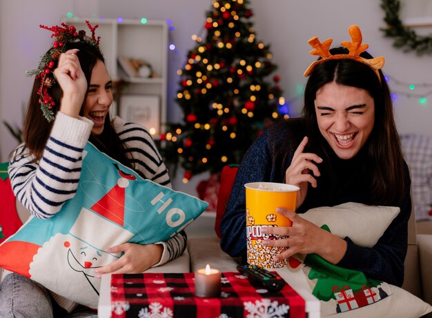 radosne, ładne młode dziewczyny z wieńcem ostrokrzewu i opaską renifera jedzą popcorn i bawią się, siedząc na fotelach i ciesząc się świętami w domu