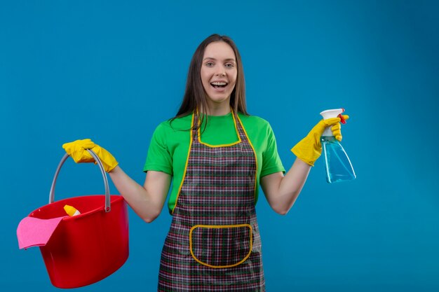 Radosne sprzątanie młoda kobieta ubrana w mundur w rękawiczkach, trzymając narzędzia do czyszczenia i spray na odizolowanej niebieskiej ścianie