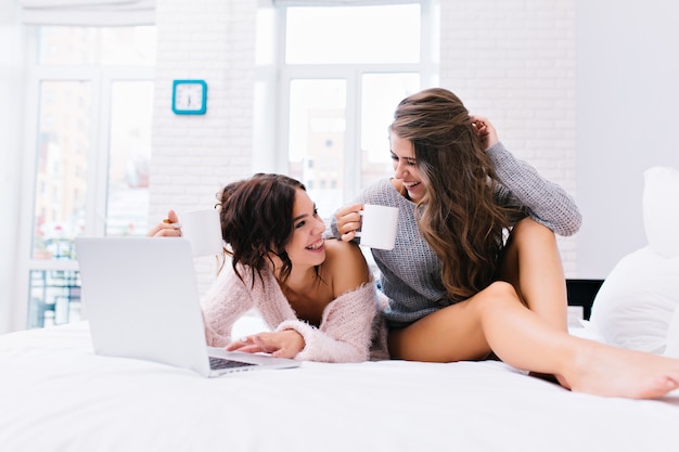 Radosny czas relaksu razem dwóch młodych atrakcyjnych kobiet zabawy na białym łóżku. Piękne modelki w wełnianych swetrach z nagimi nogami, piją herbatę, surfują po internecie, cieszą się porankiem.