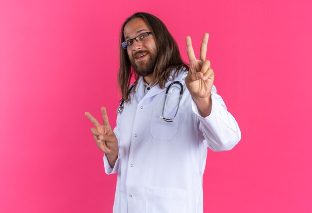 Radosny dorosły mężczyzna lekarz ubrany w szatę medyczną i stetoskop w okularach stojących w widoku profilu, robiący znak pokoju patrząc na kamerę odizolowaną na różowej ścianie