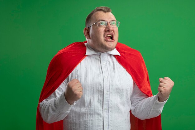 Radosny dorosły słowiański człowiek superbohatera w czerwonej pelerynie w okularach, patrząc w górę, robi gest tak na białym tle na zielonej ścianie