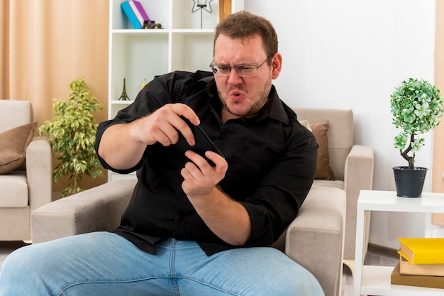 Bezpłatne zdjęcie radosny dorosły słowiański mężczyzna w okularach optycznych siedzi na fotelu grając na telefonie w salonie