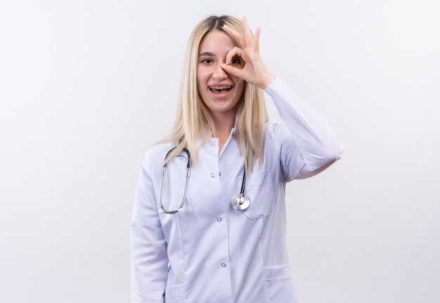 Radosny lekarz młoda blondynka ubrana w stetoskop i fartuch medyczny w ortezie dentystycznej pokazujący gest wyglądu na odizolowanej białej ścianie