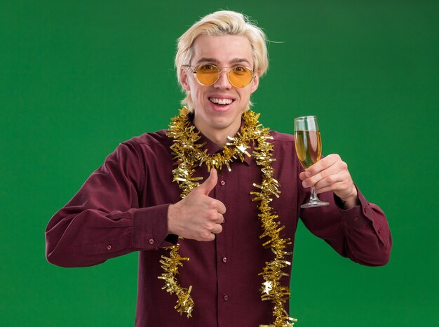 Radosny młody blondyn w okularach z świecącą girlandą na szyi trzymający kieliszek szampana pokazujący kciuk w górę odizolowany na zielonej ścianie