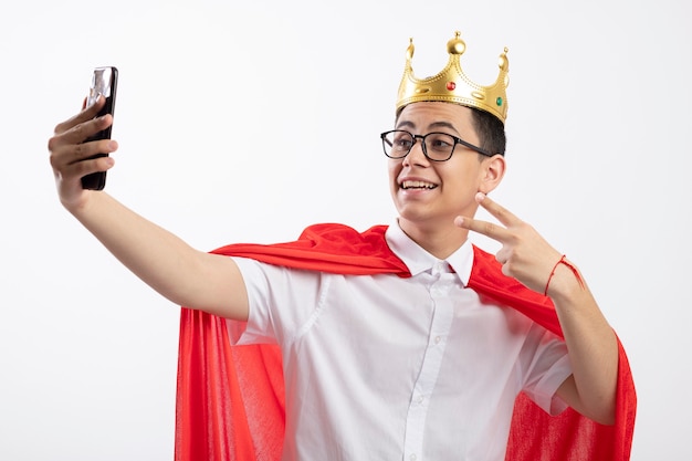 Radosny młody chłopak superbohatera w czerwonej pelerynie w okularach i koronie robi znak pokoju biorąc selfie na białym tle