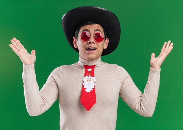 Radosny młody chłopak w kapeluszu z krawatem Boże Narodzenie i okulary, rozkładając ręce na białym tle na zielonej ścianie