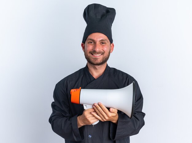 Radosny młody kucharz kaukaski w mundurze szefa kuchni i czapce trzymającej głośnik patrzący na kamerę odizolowaną na białej ścianie