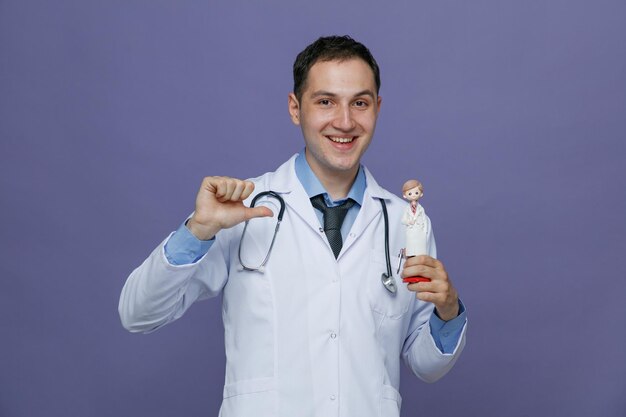 Radosny młody lekarz mężczyzna ubrany w szatę medyczną i stetoskop na szyi pokazujący figurkę lekarza wskazującą na to patrząc na kamerę odizolowaną na fioletowym tle