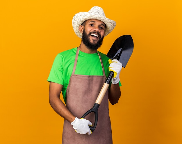 Radosny młody ogrodnik afro-amerykański facet ubrany ogrodniczy kapelusz i rękawiczki, trzymający łopatę na białym tle na pomarańczowej ścianie