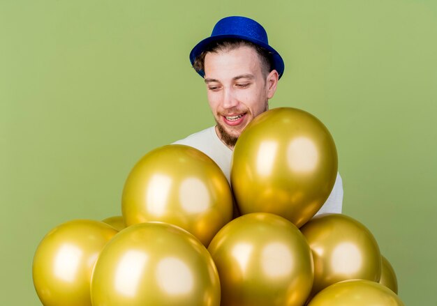 Radosny młody przystojny facet słowiańskich partii na sobie kapelusz partii stojącej za balonami i patrząc na nich na białym tle na oliwkowym tle