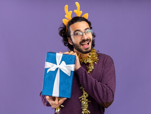 Radosny młody przystojny facet ubrany w Boże Narodzenie obręcz do włosów z girlandą na szyi, trzymając pudełko na białym tle na niebieskiej ścianie