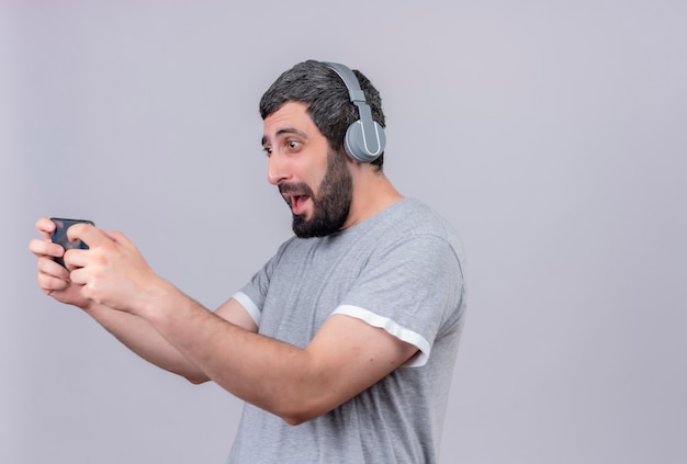 Radosny młody przystojny kaukaski mężczyzna nosi słuchawki za pomocą swojego telefonu komórkowego na białym tle