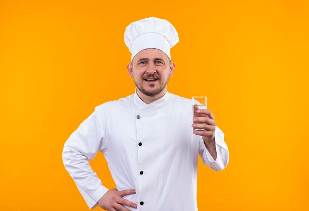 Radosny młody przystojny kucharz w mundurze szefa kuchni trzymający szklankę wody odizolowaną na pomarańczowej ścianie