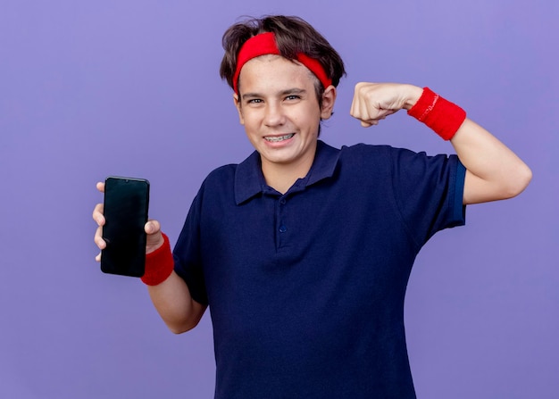 Radosny młody przystojny sportowy chłopiec noszący opaskę i opaski na nadgarstki z szelkami dentystycznymi pokazujący telefon komórkowy robi silny gest patrząc z przodu odizolowany na fioletowej ścianie
