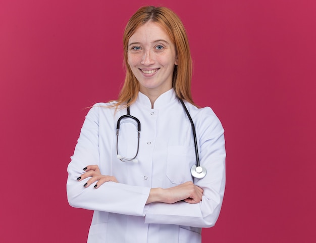 Radosny młody żeński imbirowy lekarz ubrany w szatę medyczną i stetoskop stojący z zamkniętą postawą odizolowaną na szkarłatnej ścianie z kopią przestrzeni
