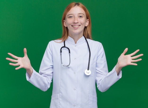 Bezpłatne zdjęcie radosny młody żeński lekarz imbir nosi szatę medyczną i stetoskop pokazując puste ręce izolowane na zielonej ścianie