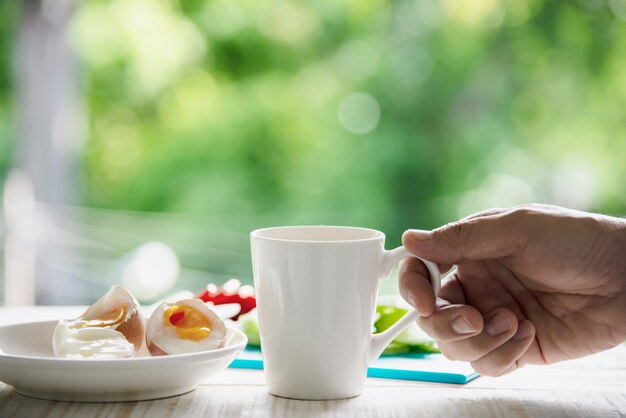 Ręczne kapturowanie gorącej filiżanki kawy z gotowanymi jajkami ze świeżą ogórkiem sałatka ziemniaczana cebula śniadanie zestaw z zielonym lesie - koncepcja żywności śniadanie