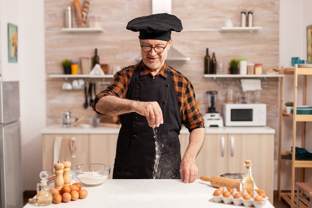 Ręczne rozprowadzanie mąki pszennej na drewnianym stole w kuchni do domowej pizzy. Emerytowany starszy kucharz z bonete i fartuchem, w mundurze kuchennym zraszającym ręcznie przesiewającym składniki.