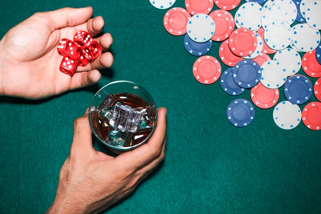Ręka mężczyzny trzyma czerwone kostki i szkła whisky nad stołem do pokera