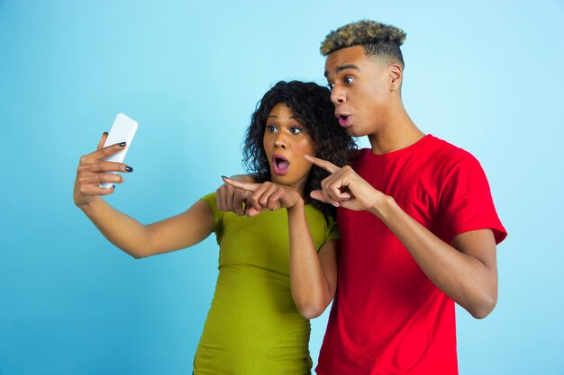 Robienie razem selfie lub vloga. Młody emocjonalny Afroamerykanin mężczyzna i kobieta w kolorowe ubrania na niebieskim tle. Piękna para. Pojęcie ludzkich emocji, wyraz twarzy, relacje, reklama.