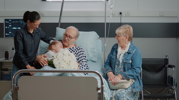 Bezpłatne zdjęcie rodzina odwiedzająca starego pacjenta i przynosząca kwiaty na szpitalnym łóżku. matka i dziecko przytulają starszego mężczyznę podczas wizyty rodzinnej w klinice, ciesząc się zdrowieniem z córką i małą dziewczynką. goście